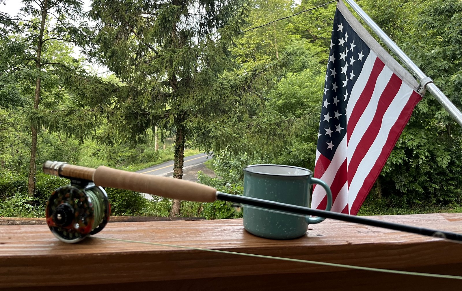 A fly rod with a hand-painted reel resting on a porch rail beside an enamel coffee mug, green woods and a flag beyond